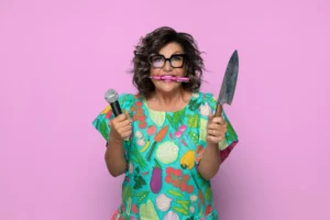 A woman holding a microphone and knife, smiling against a vibrant pink background.