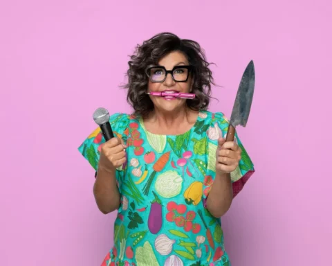 A woman holding a microphone and knife, smiling against a vibrant pink background.