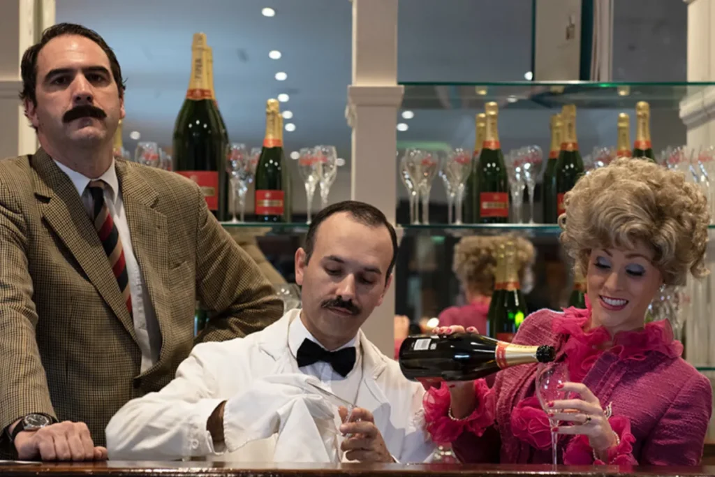 A man and woman sit at a bar, smiling and toasting with a bottle of champagne between them.