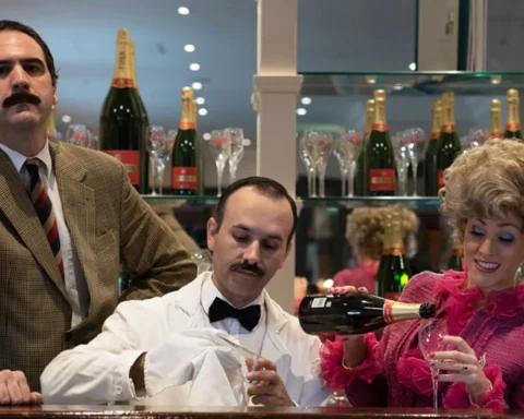 A man and woman sit at a bar, smiling and toasting with a bottle of champagne between them.