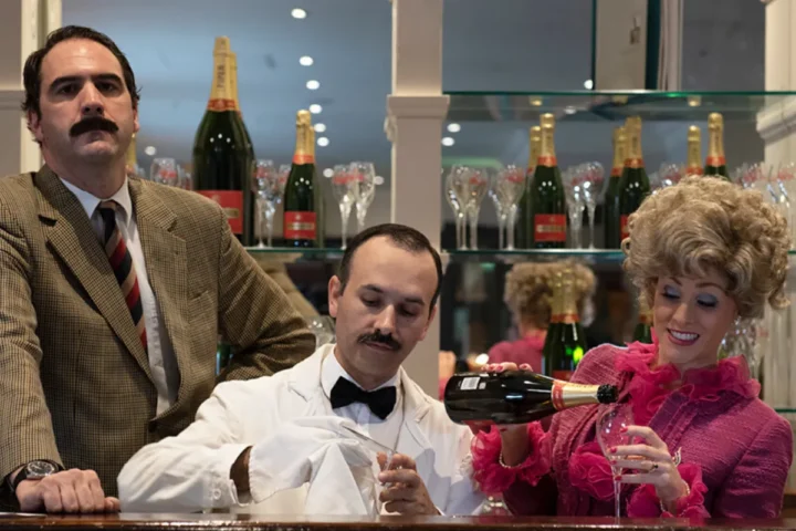A man and woman sit at a bar, smiling and toasting with a bottle of champagne between them.