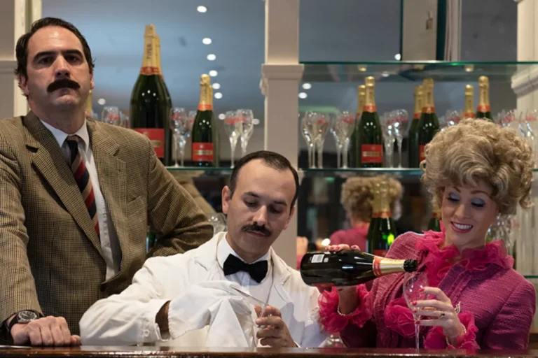 A man and woman sit at a bar, smiling and toasting with a bottle of champagne between them.