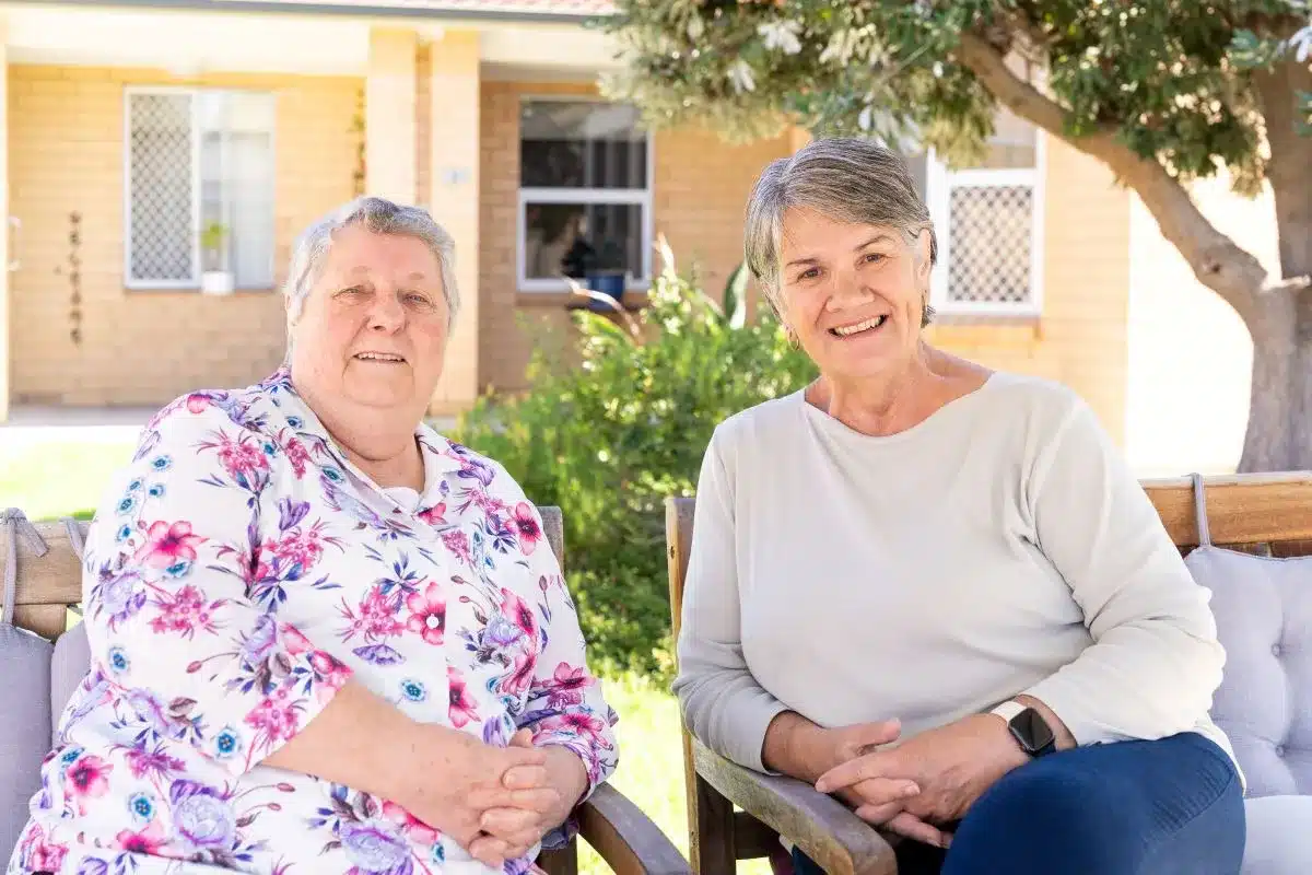 Two women sit on a bench outside a house, enjoying a conversation in a sunny outdoor setting.