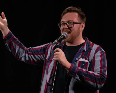 A man wearing glasses and a plaid shirt holds a microphone, appearing engaged in a speaking or performance activity.