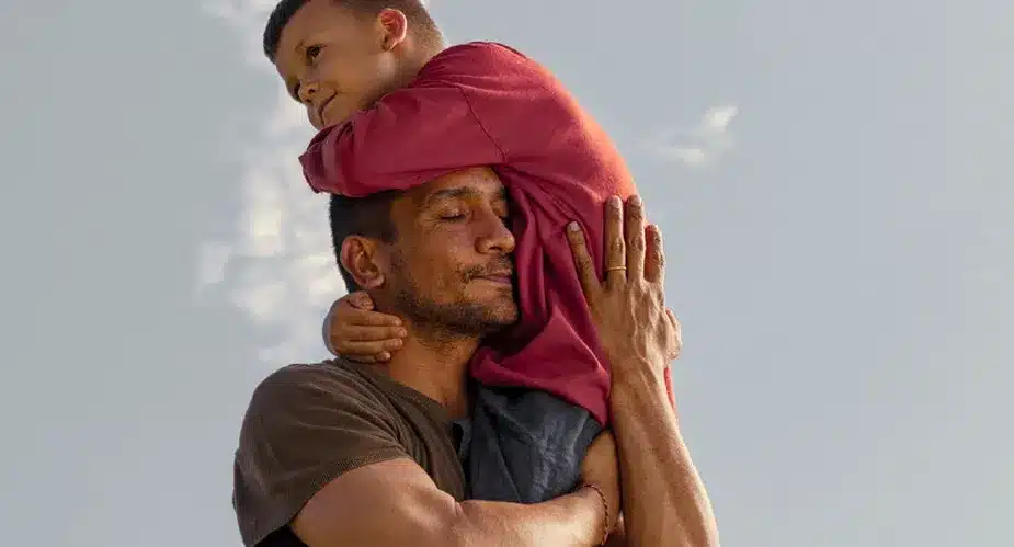 A man carries a child on his shoulders, both smiling and enjoying a sunny day outdoors.