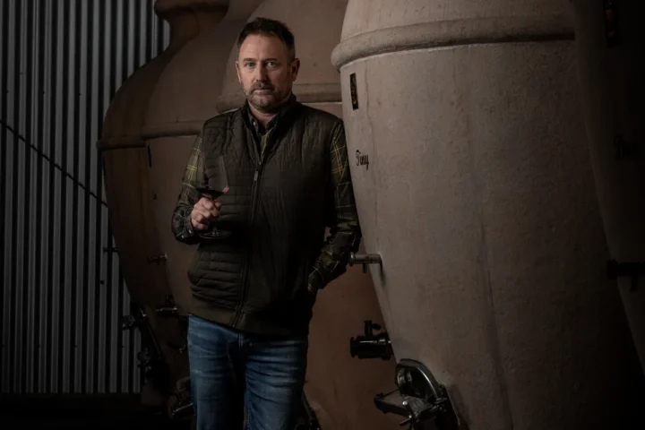 A man stands beside a large tank surrounded by stacked wine barrels in a winery setting.