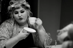 A woman holds a curling iron in one hand and a cup of coffee in the other, preparing for her hairstyle.