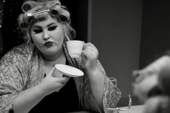 A woman holds a curling iron in one hand and a cup of coffee in the other, preparing for her hairstyle.