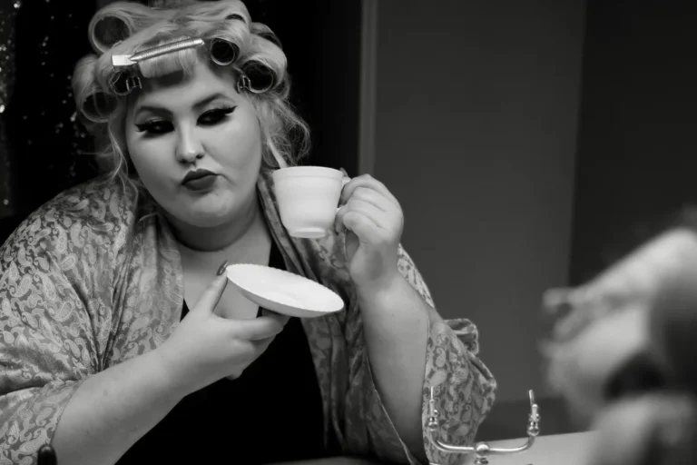 A woman holds a curling iron in one hand and a cup of coffee in the other, preparing for her hairstyle.
