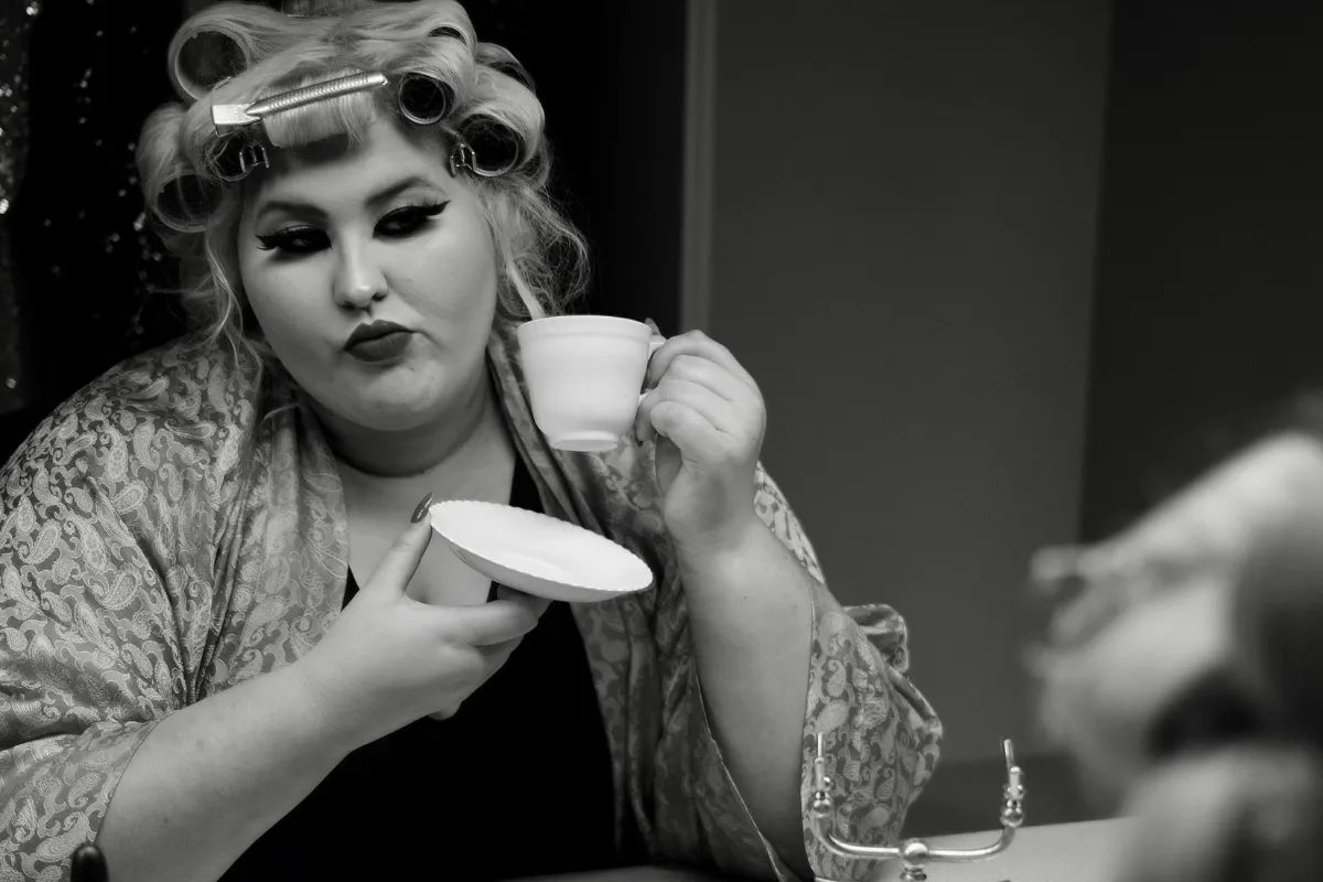 A woman holds a curling iron in one hand and a cup of coffee in the other, preparing for her hairstyle.