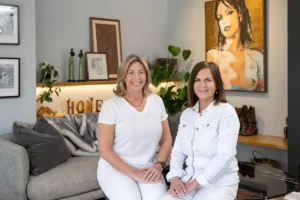 Two women relax on a couch, chatting in front of a colorful painting hanging on the wall.