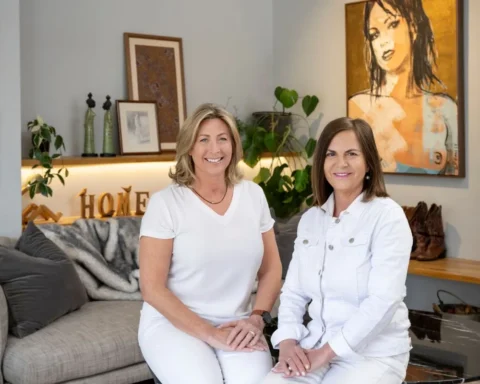 Two women relax on a couch, chatting in front of a colorful painting hanging on the wall.