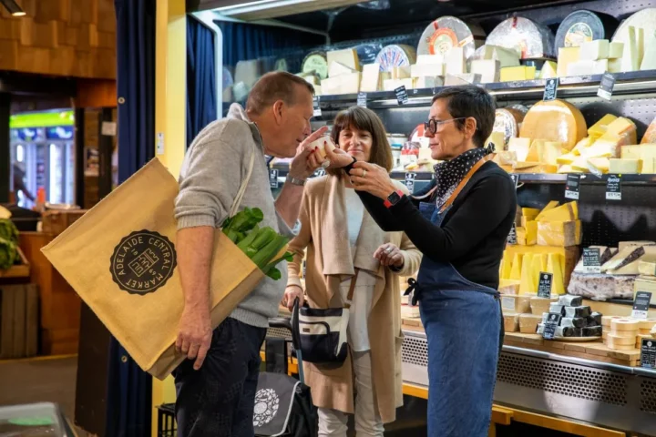 A man and woman stand together, smiling in front of a colorful cheese display at a market.