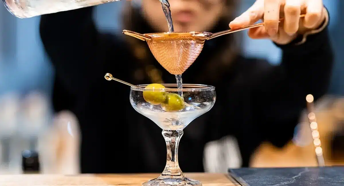 A woman elegantly pours a martini from a shaker into a glass, showcasing her bartending skills.