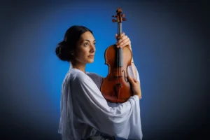 A woman in white attire holds a violin, poised and ready to play.