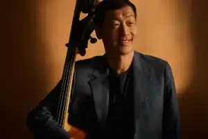 A man in a suit stands holding a cello, showcasing a blend of elegance and musical artistry.