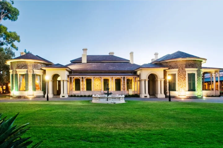 A large mansion's front view featuring an elegant fountain in the center of a landscaped yard.