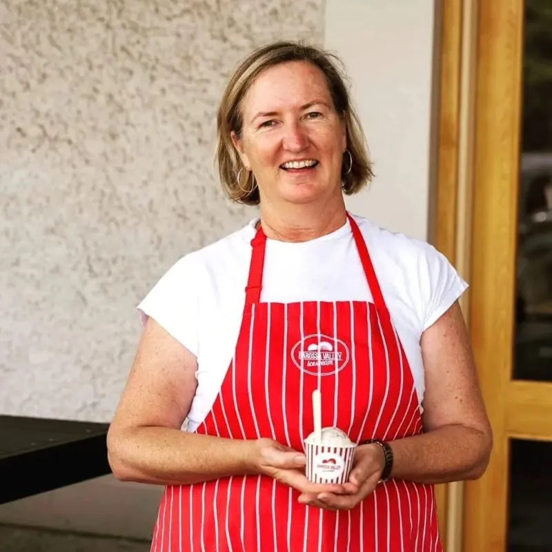 A woman in a red and white apron smiles while holding a colorful cupcake in her hand.