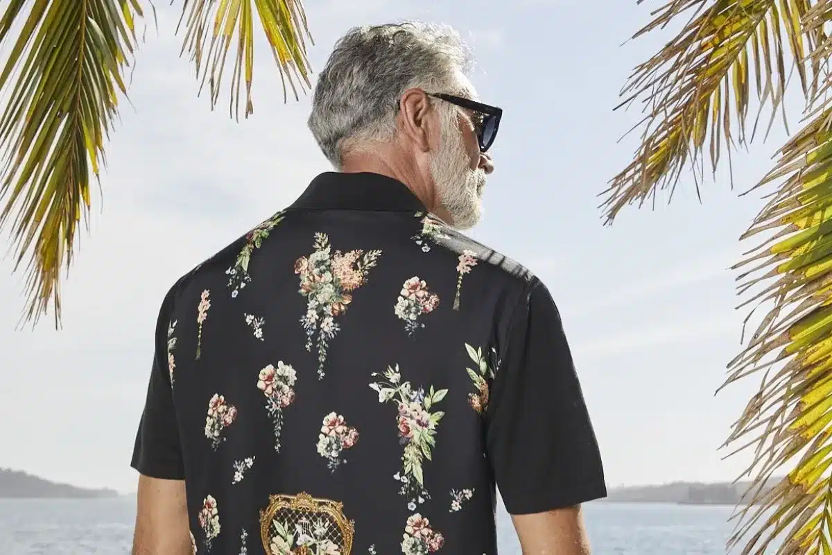 A man in sunglasses and a floral shirt gazes thoughtfully out at the ocean.