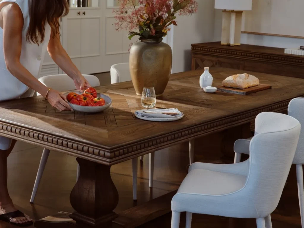 A woman stands at a table, smiling, with a colorful bowl of assorted fruit in front of her.