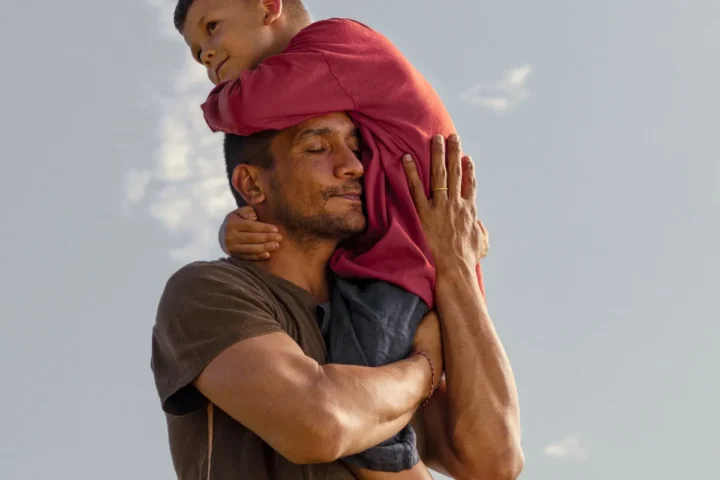 A man carries a child on his shoulders, both smiling and enjoying a sunny day outdoors.