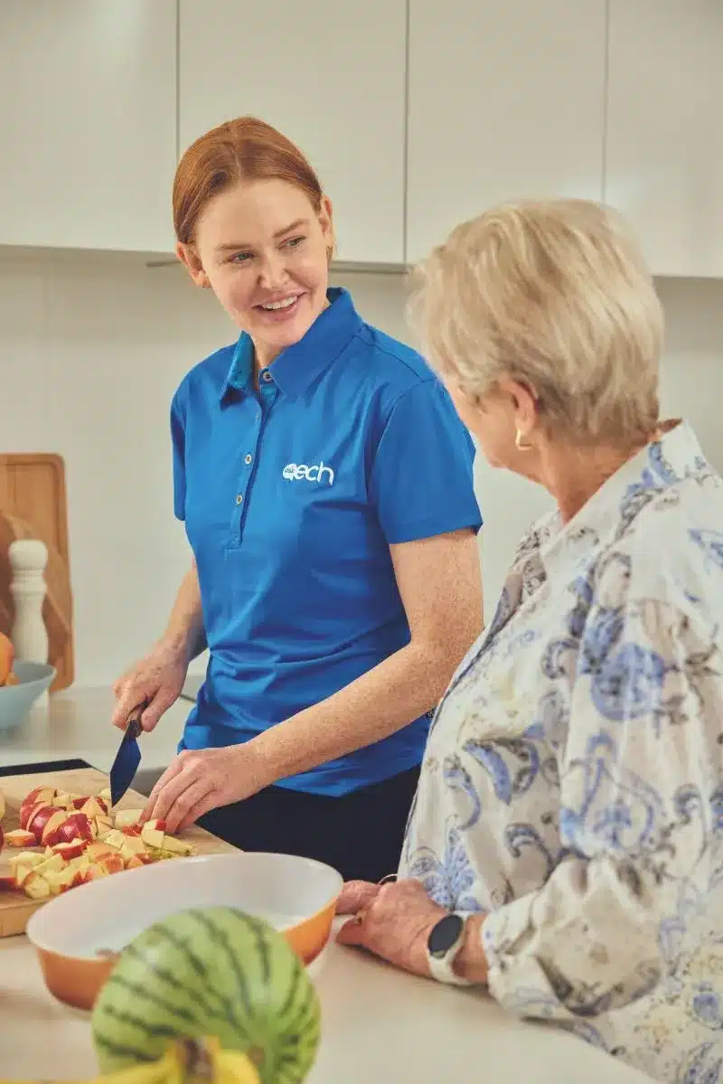 A woman in a blue shirt assists an older woman while cooking in a cozy kitchen.