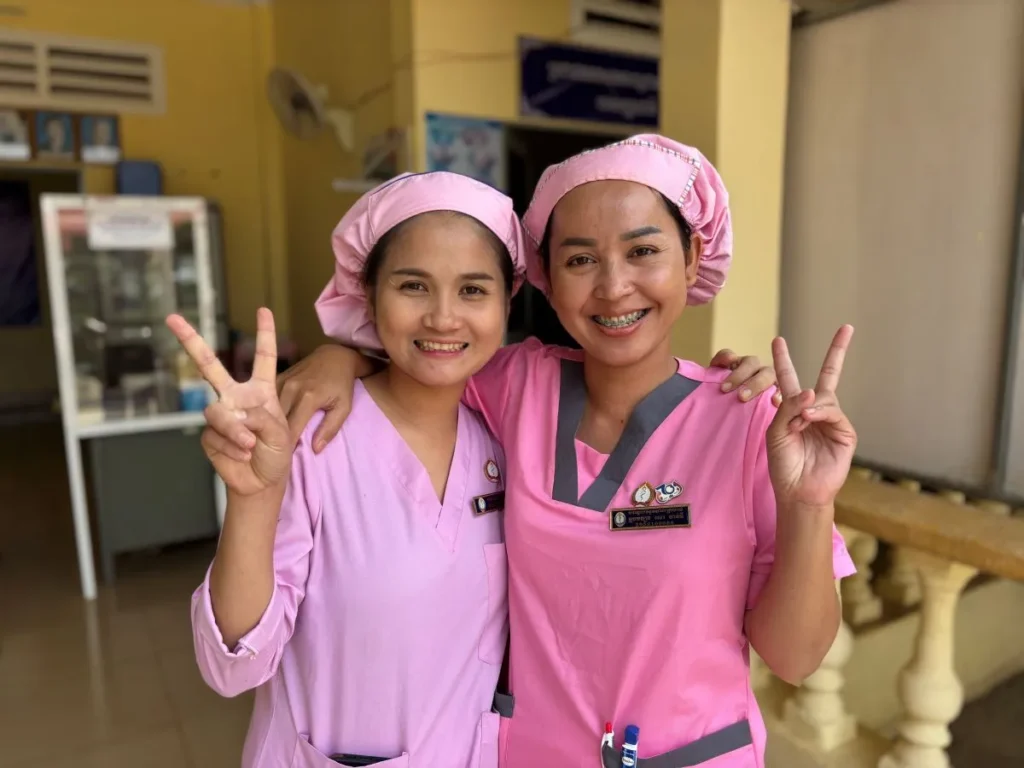 Two women in pink scrubs smile and give the peace sign with their fingers.