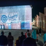 A group of people stands in front of a building displaying a colorful projection at night.