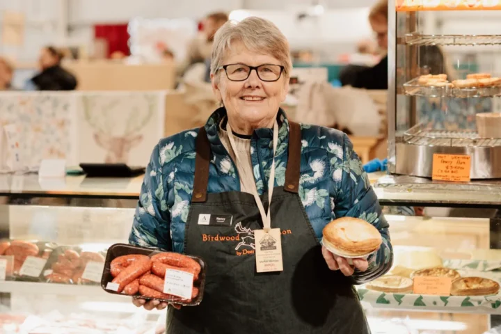 A woman in an apron holds a sandwich in one hand and a bun in the other, smiling at the camera.