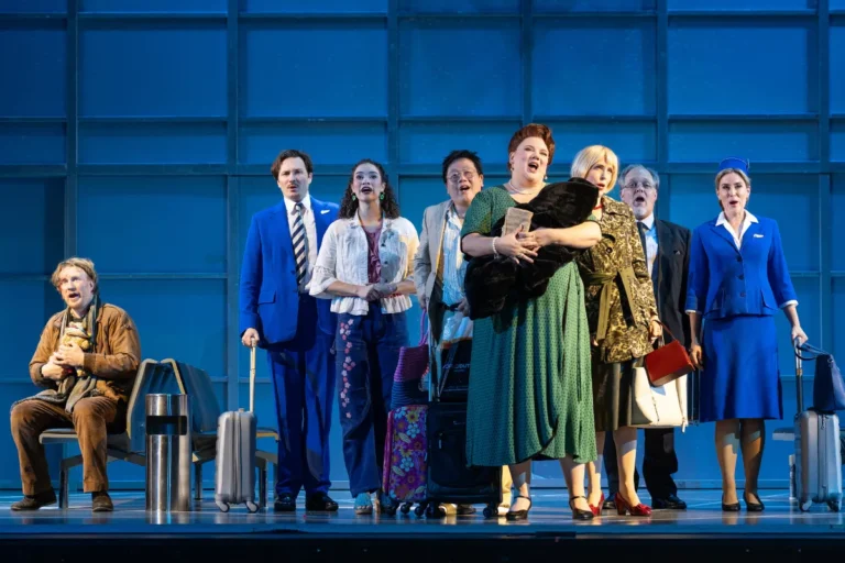 A group of people on stage, each holding luggage, smiling and engaging with the audience.