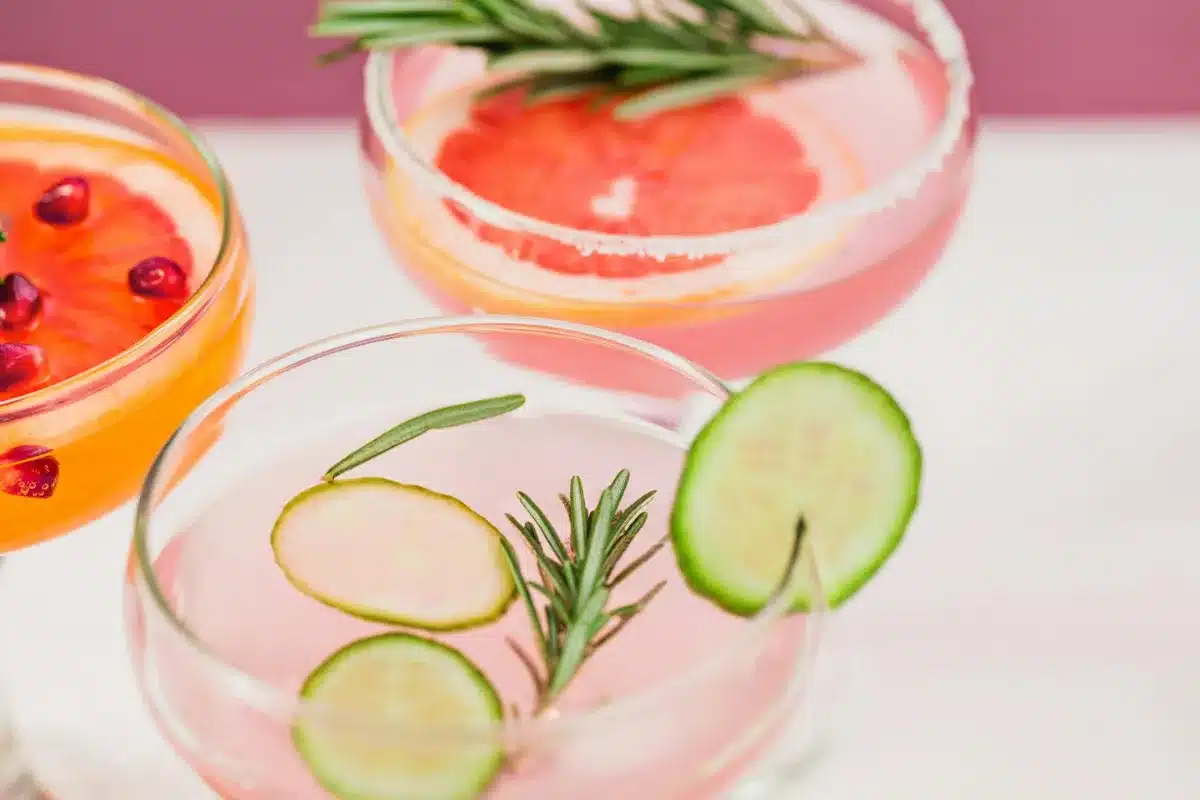 Three glasses of pink drinks garnished with cucumber, rosemary, and lemon slices on a wooden table.