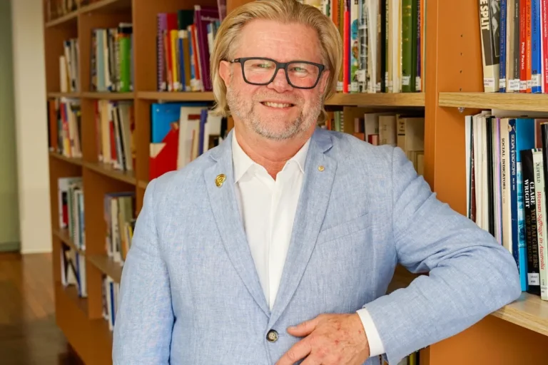 A man wearing glasses and a blue blazer stands in front of tall bookshelves filled with books.