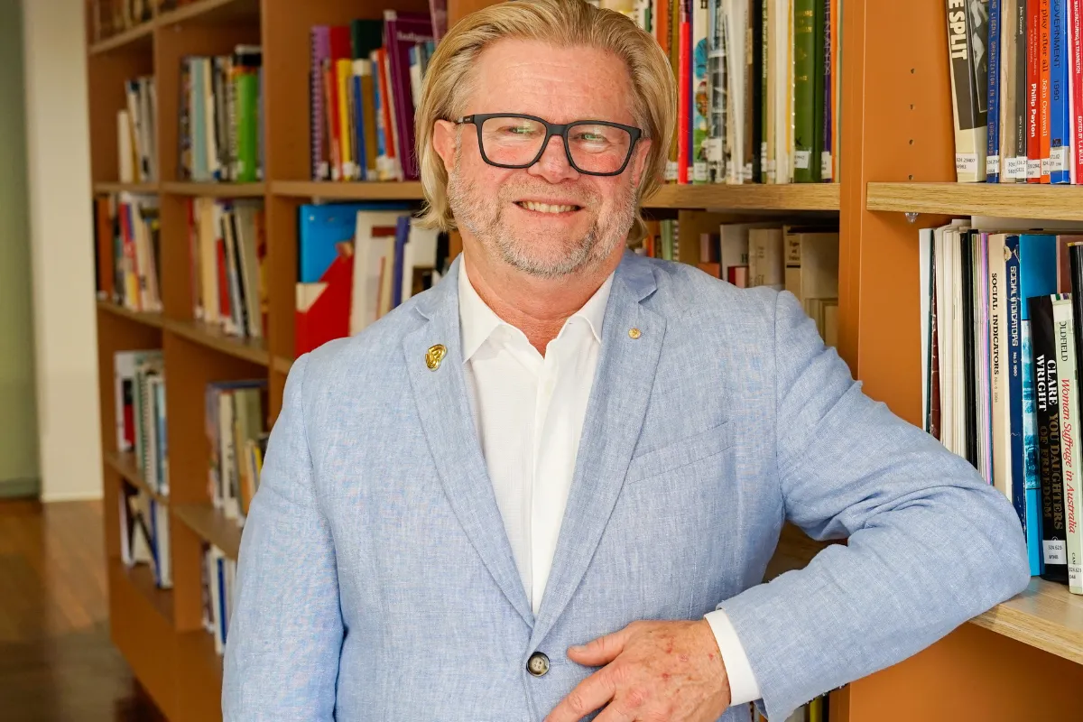 A man wearing glasses and a blue blazer stands in front of tall bookshelves filled with books.