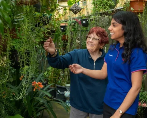 A woman and an older woman smiling together in a vibrant garden filled with flowers and greenery.