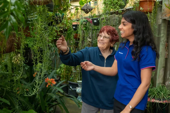 A woman and an older woman smiling together in a vibrant garden filled with flowers and greenery.