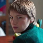 A woman wearing a green sweater sits at a table, looking thoughtfully at something in front of her.