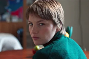 A woman wearing a green sweater sits at a table, looking thoughtfully at something in front of her.
