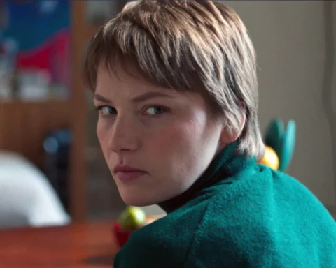 A woman wearing a green sweater sits at a table, looking thoughtfully at something in front of her.
