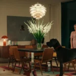 A man and woman stand in a cozy living room, smiling at each other with a dog sitting at their feet.