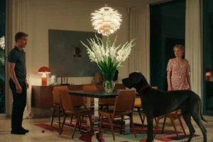 A man and woman stand in a cozy living room, smiling at each other with a dog sitting at their feet.