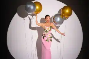 A woman in a pink dress joyfully holds colorful balloons against a bright background.