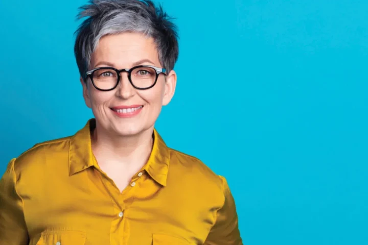 A woman wearing glasses and a bright yellow shirt smiles at the camera.