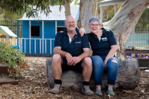 An older couple sits on a rock, smiling, with a colorful playground in the background.