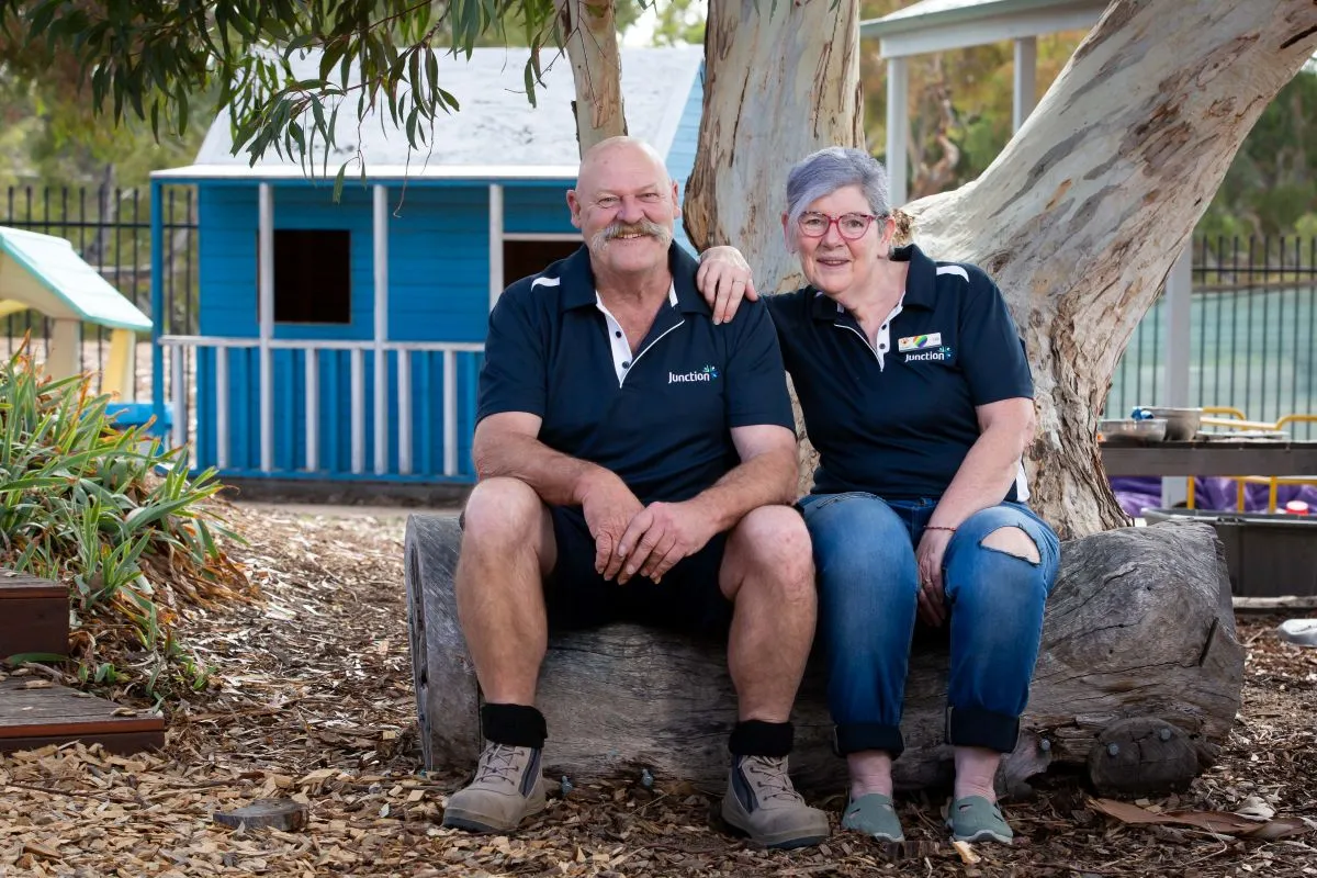 An older couple sits on a rock, smiling, with a colorful playground in the background.