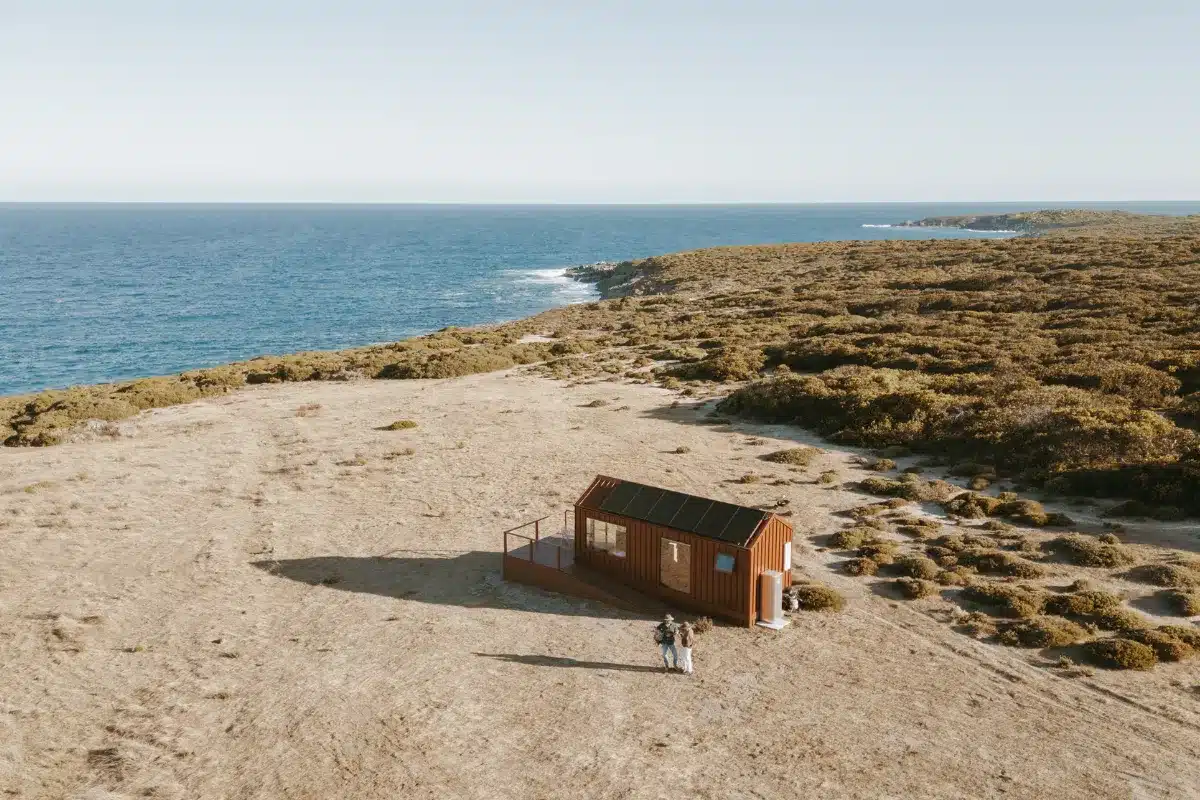A small beach cabin sits near the ocean, surrounded by sand and gentle waves under a clear blue sky.