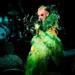 A person in a vibrant green feather costume passionately sings into a microphone on stage.
