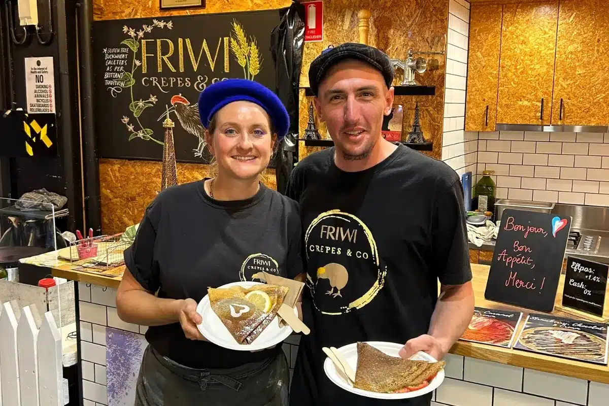 Two people stand in front of a counter, each holding a plate of food, smiling and engaging with each other.