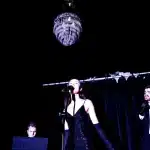A man and woman in black dresses stand beside an elegant chandelier, showcasing a sophisticated atmosphere.