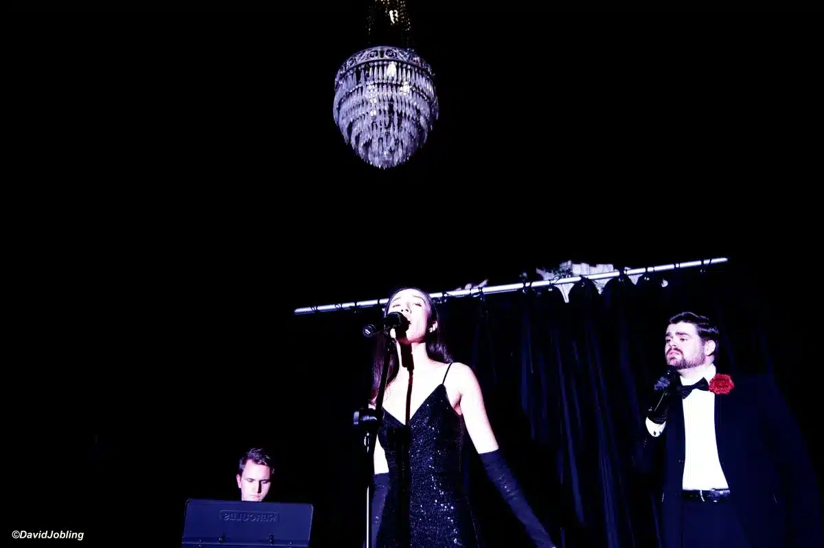 A man and woman in black dresses stand beside an elegant chandelier, showcasing a sophisticated atmosphere.