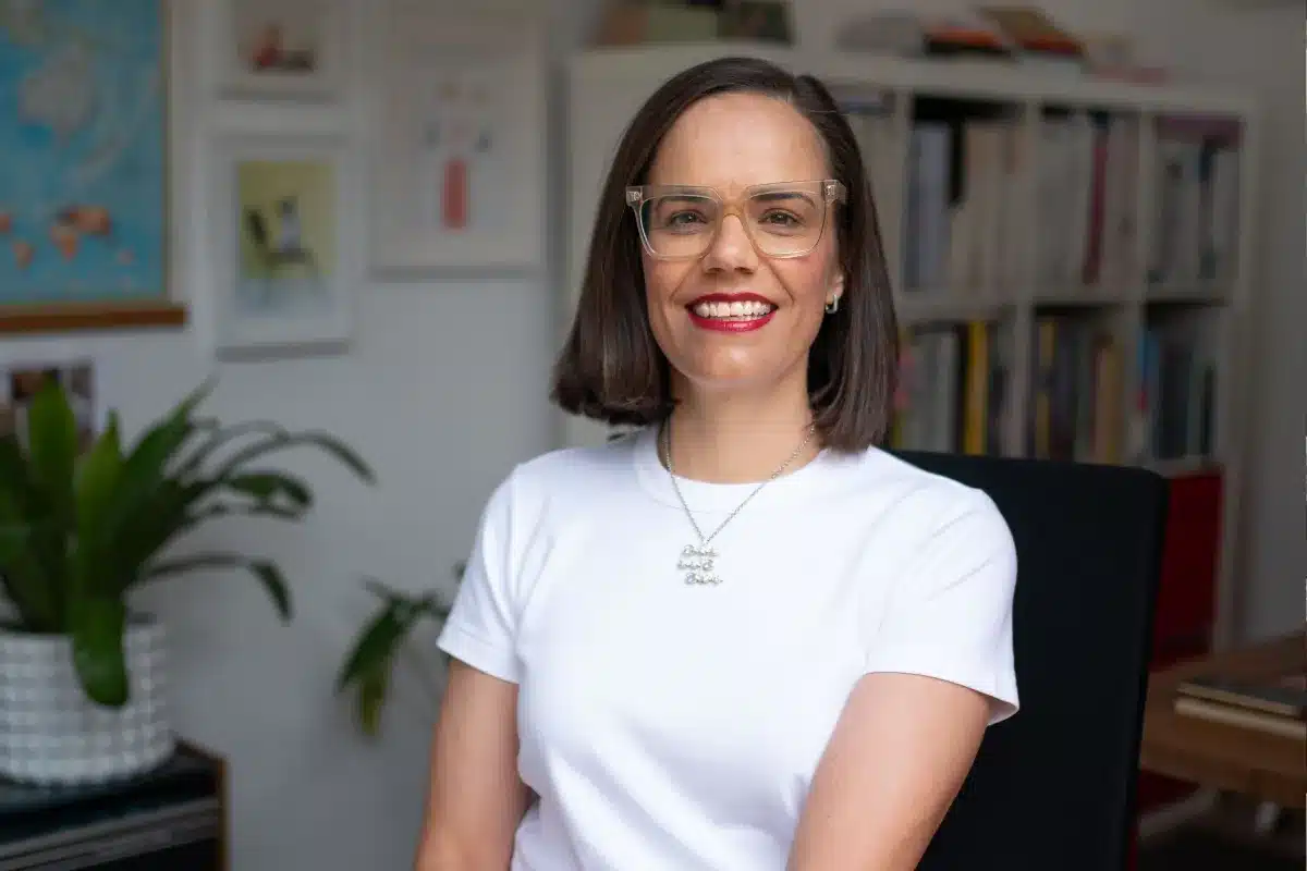 A woman wearing glasses and a white shirt sits in front of a bookcase filled with books.